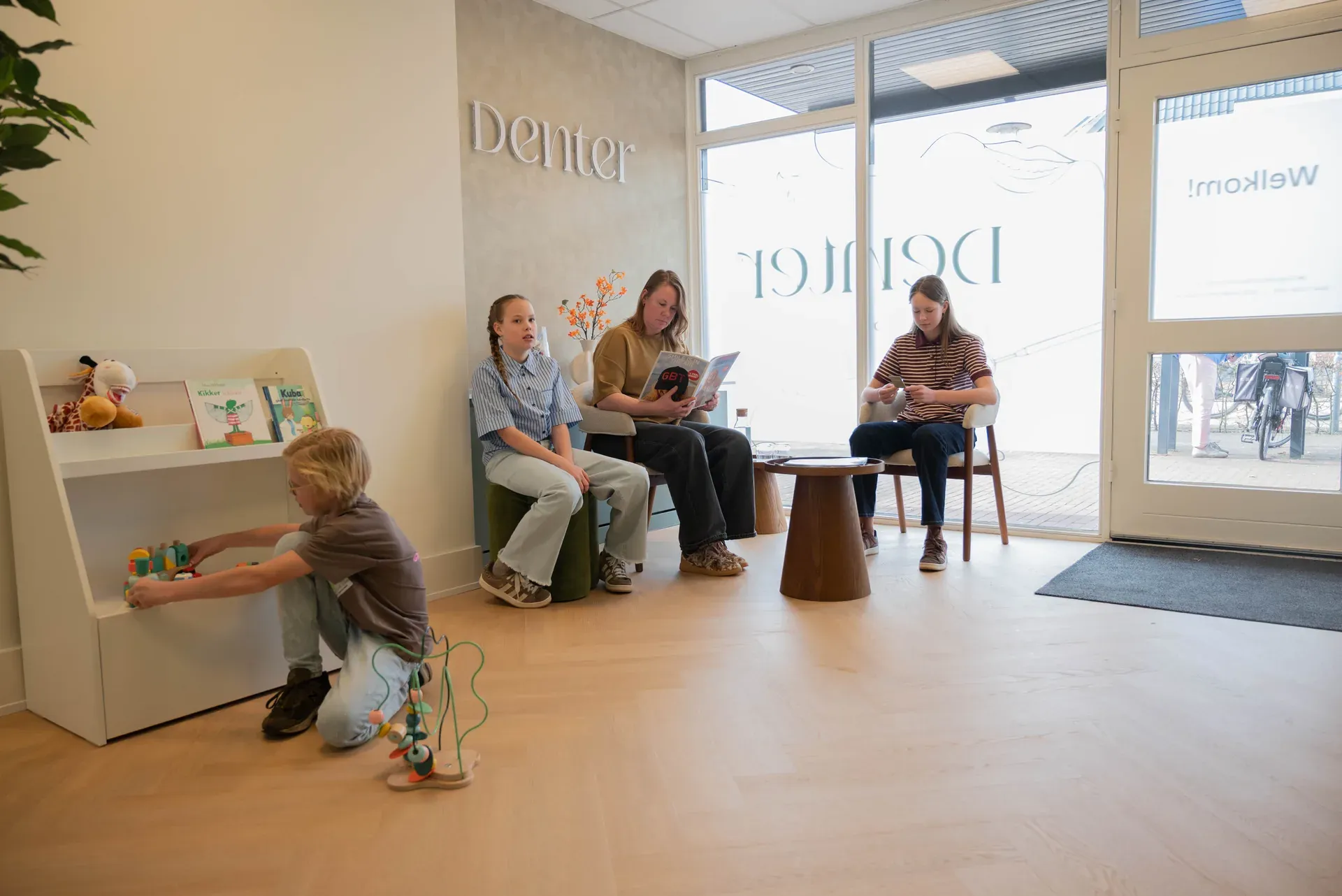 Een gezin wacht in de wachtkamer; een jongen speelt bij de boekenkast in de kinderhoek